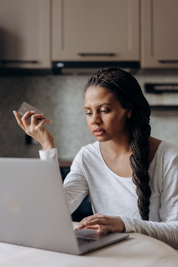 Young black woman with braided hair uses laptop and credit card for online banking in a modern home setting.